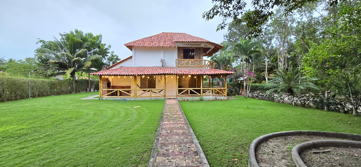 The two-story house features a terracotta tiled roof and is surrounded by a well-maintained green lawn. A stone pathway leads to the entrance, framed by tropical foliage and palm trees. The patio includes hanging hammocks for relaxation.