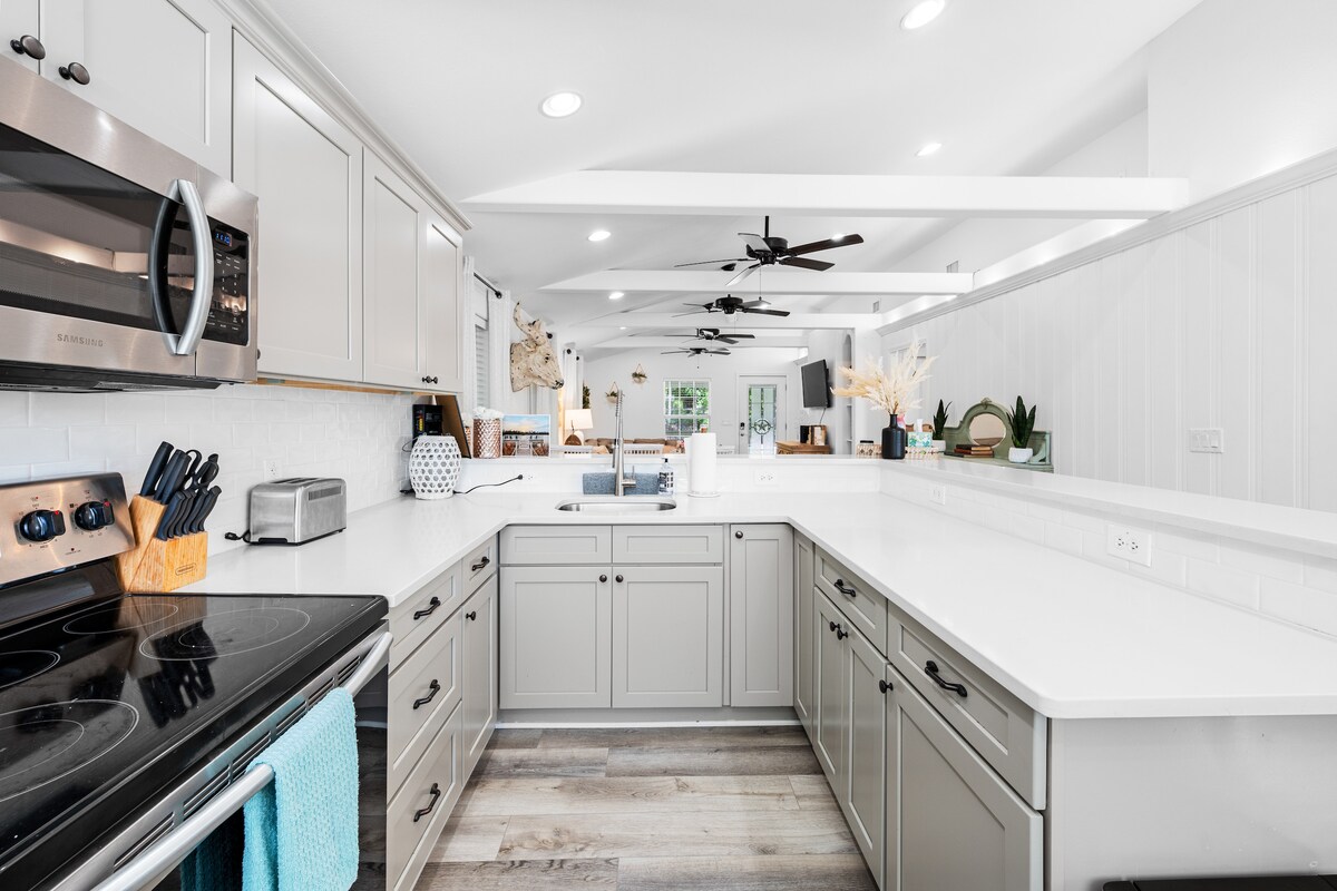 A modern kitchen features light grey cabinetry and sleek white countertops, enhancing functionality and spaciousness. Stainless steel appliances, including a microwave and stove, are visible. Illumination is provided by recessed lights, and a casual dining area can be seen in the background.