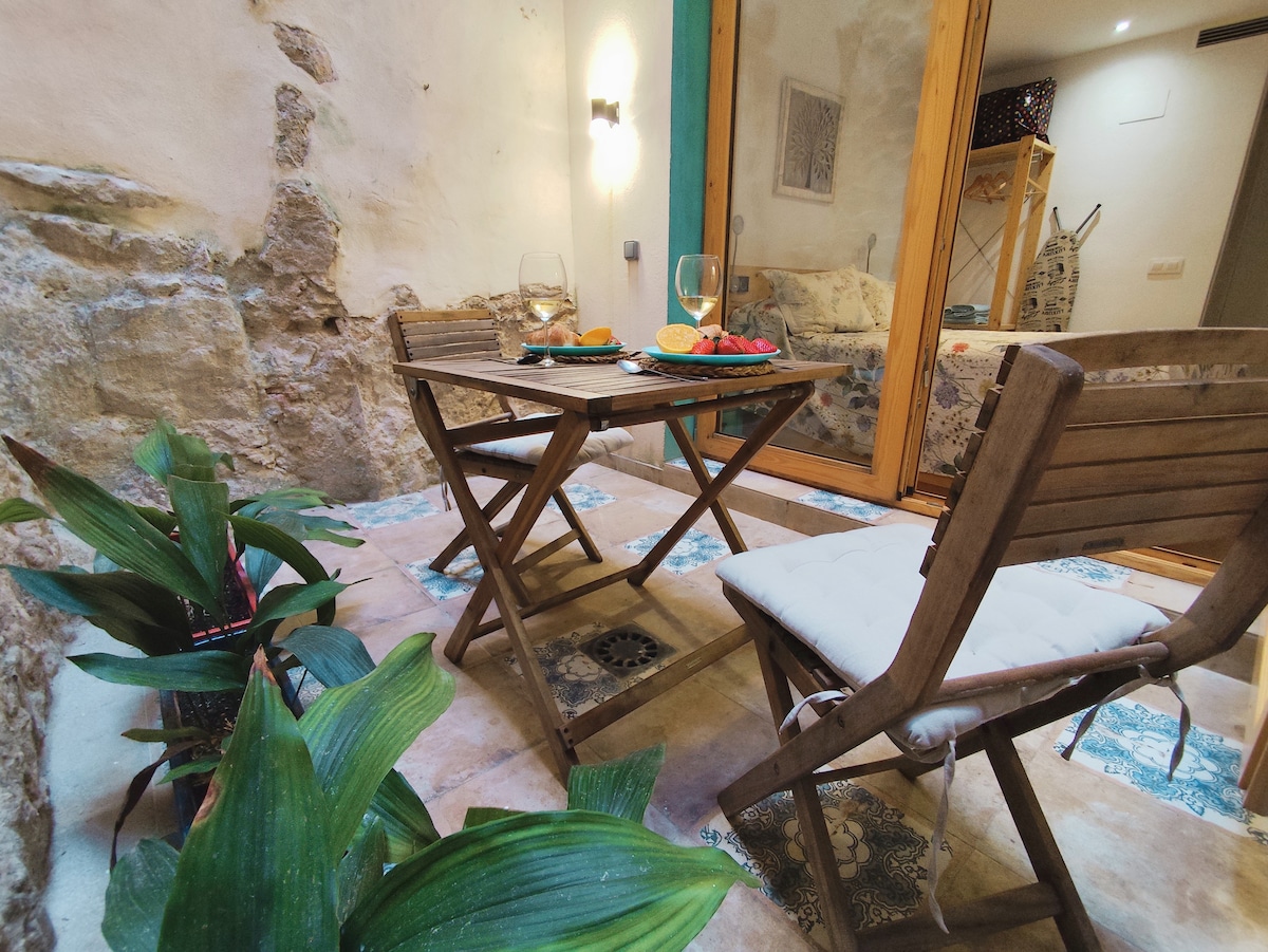 A cozy patio area features a wooden table set for two, adorned with a selection of fruits. Two folding chairs provide seating, surrounded by potted plants that enhance the tranquil atmosphere. A stone wall serves as a natural backdrop, adding charm to the space.