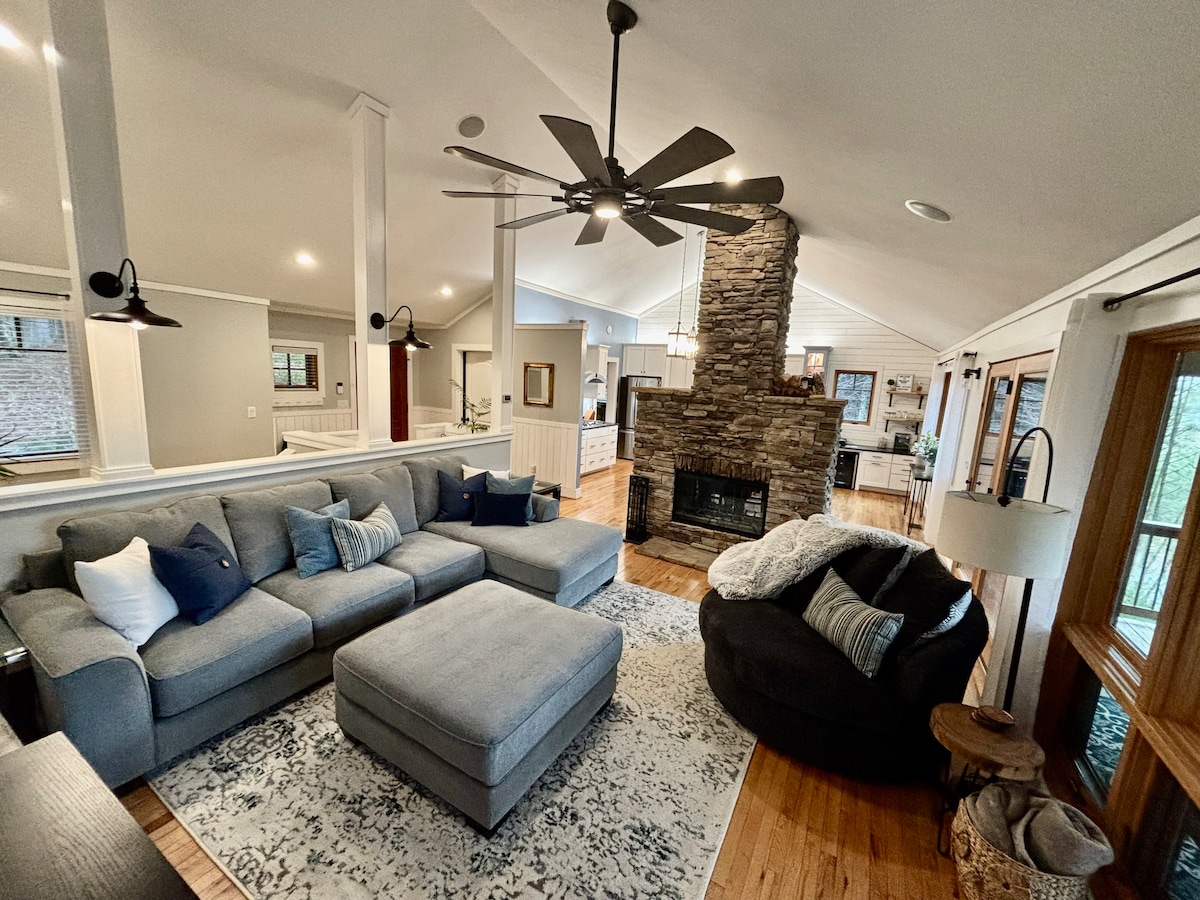 A spacious living area features a large gray sectional sofa, complemented by a matching ottoman. A textured rug lies beneath, while a stone fireplace serves as a focal point. Large windows allow natural light to brighten the space, creating a comfortable gathering spot.