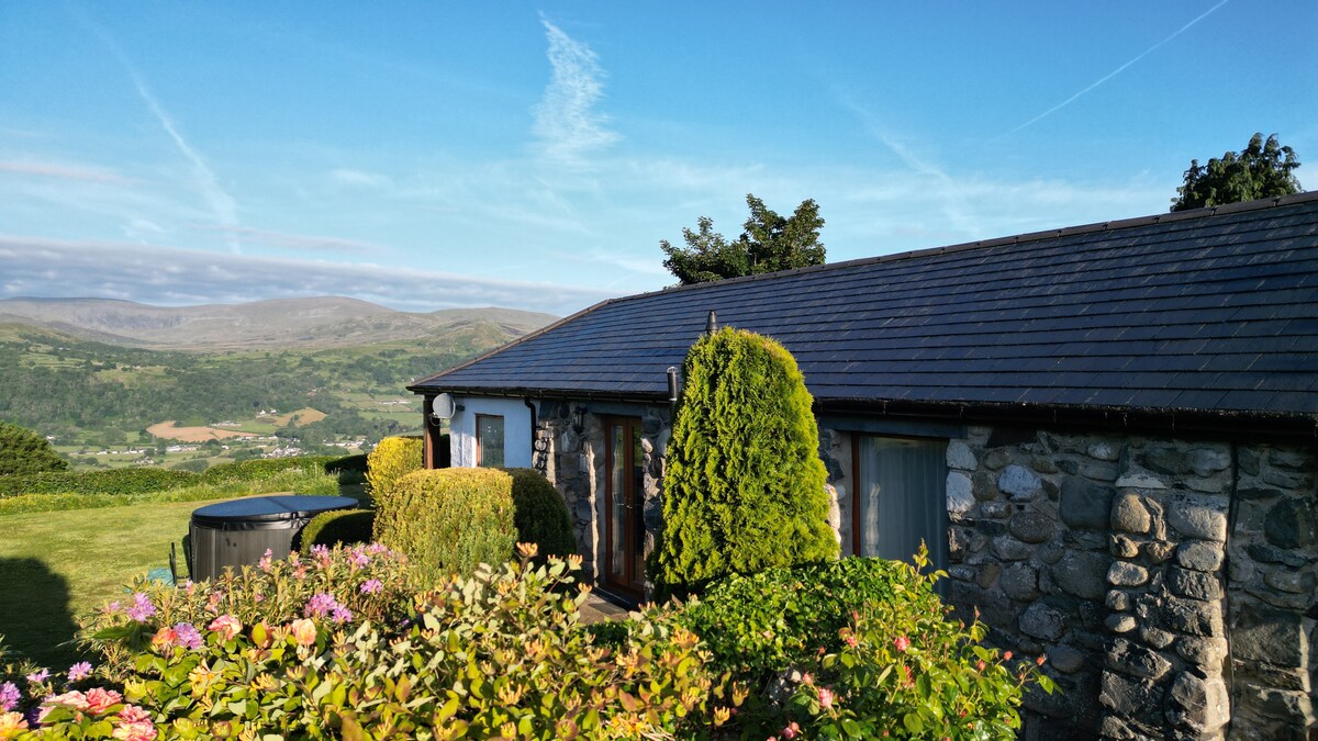 The charming stone cottage is surrounded by vibrant greenery and blooming flowers. A private hot tub is positioned on the patio, with expansive views of the valley and Snowdonia's mountains visible in the background under a clear blue sky.