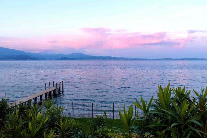 A serene view of Lake Garda is presented, featuring calm waters under a pastel sky. A wooden dock extends into the lake, surrounded by lush greenery. The landscape includes distant mountains, creating a peaceful backdrop for the scene.