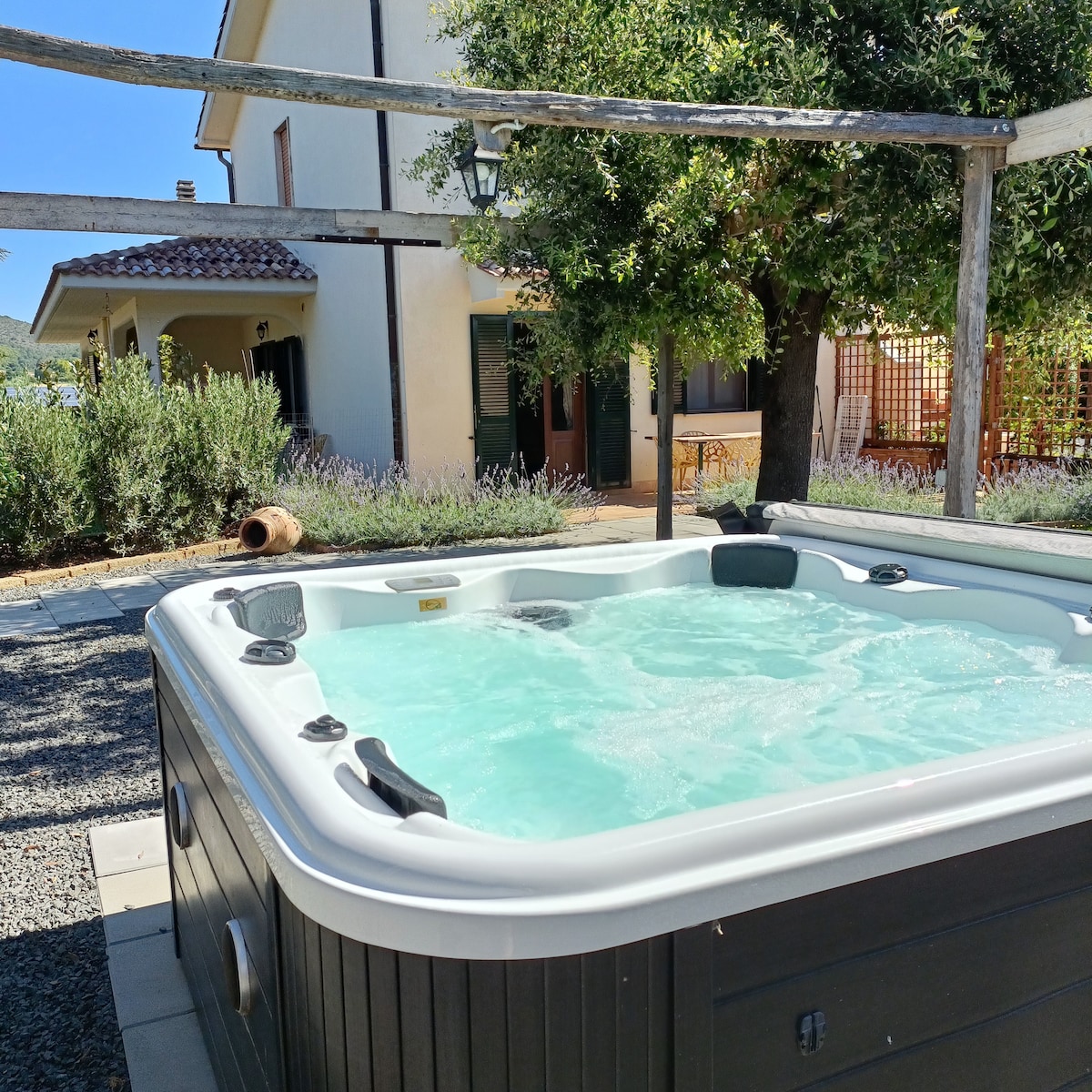 A private hot tub accommodates six people, surrounded by a stone patio. Lush greenery and blooming lavender enhance the outdoor space, while a charming house stands in the background, providing a serene setting for relaxation.