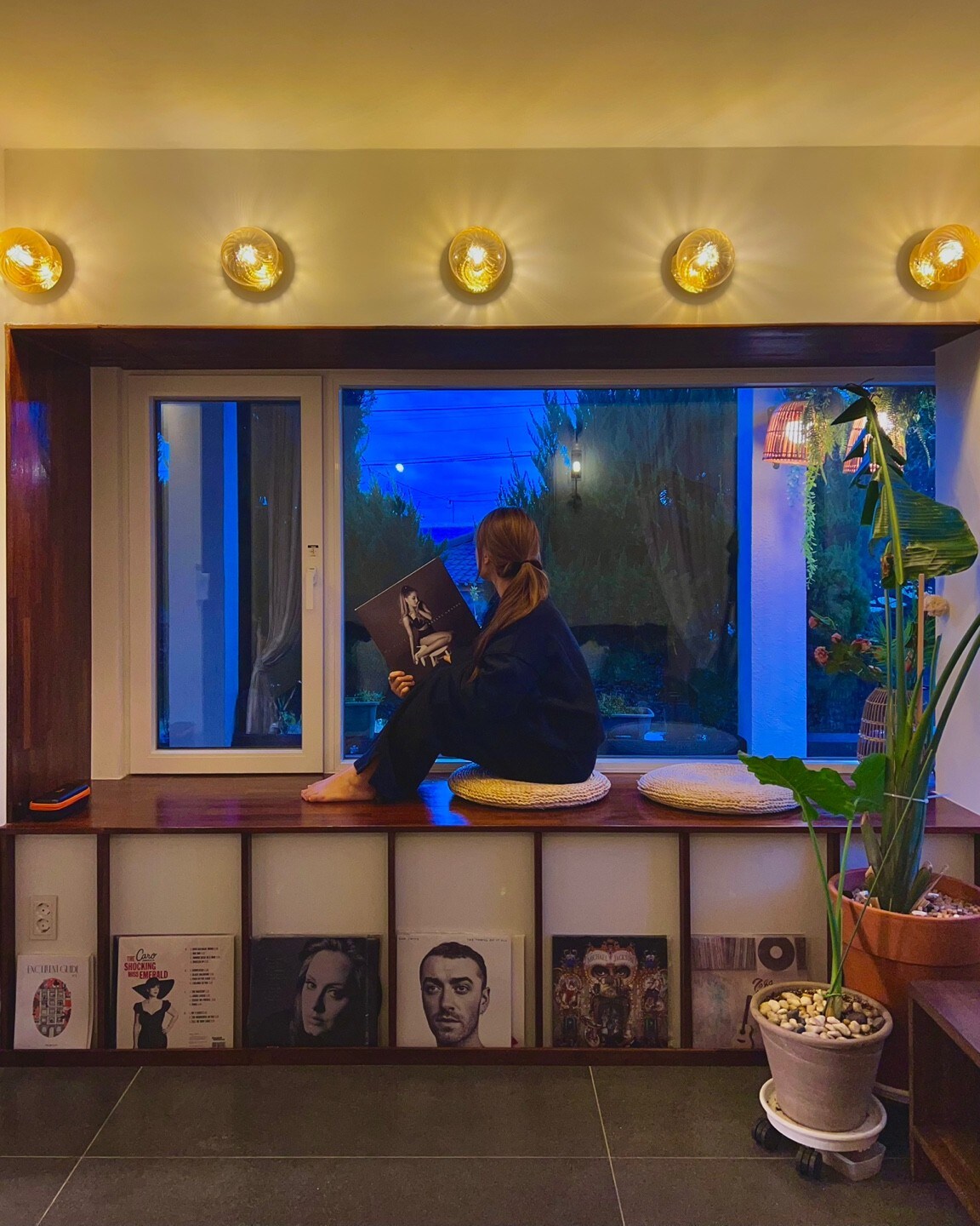 A cozy reading nook features a large window illuminated by soft lights. A person sits comfortably with their legs crossed, holding an album cover. Indoor plants and decorative stones are arranged on a nearby shelf, while books are neatly displayed beneath.