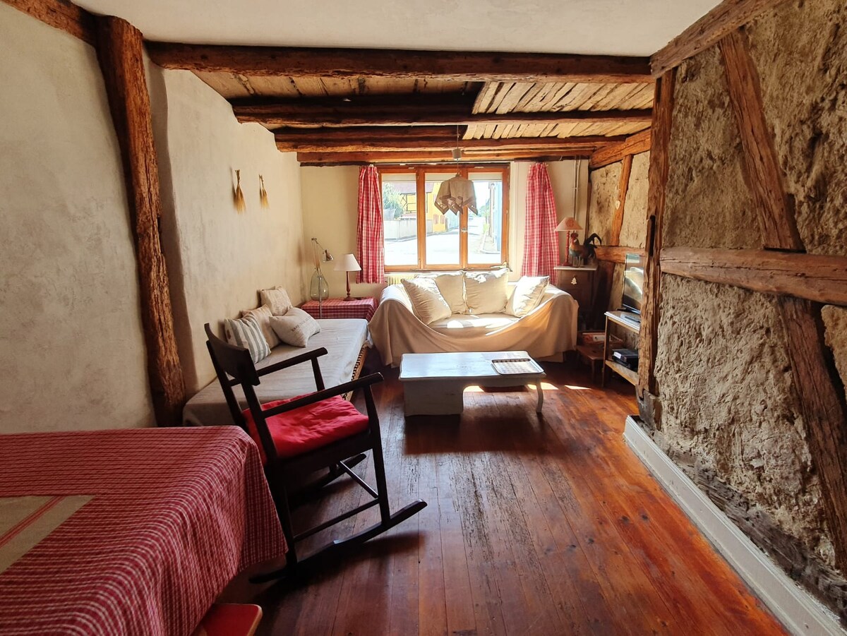 A cozy living space features wooden beams and rustic stone walls, with a white sofa and armchair arranged around a coffee table. Natural light enters through a window dressed with red curtains, enhancing the warm ambiance of the room.