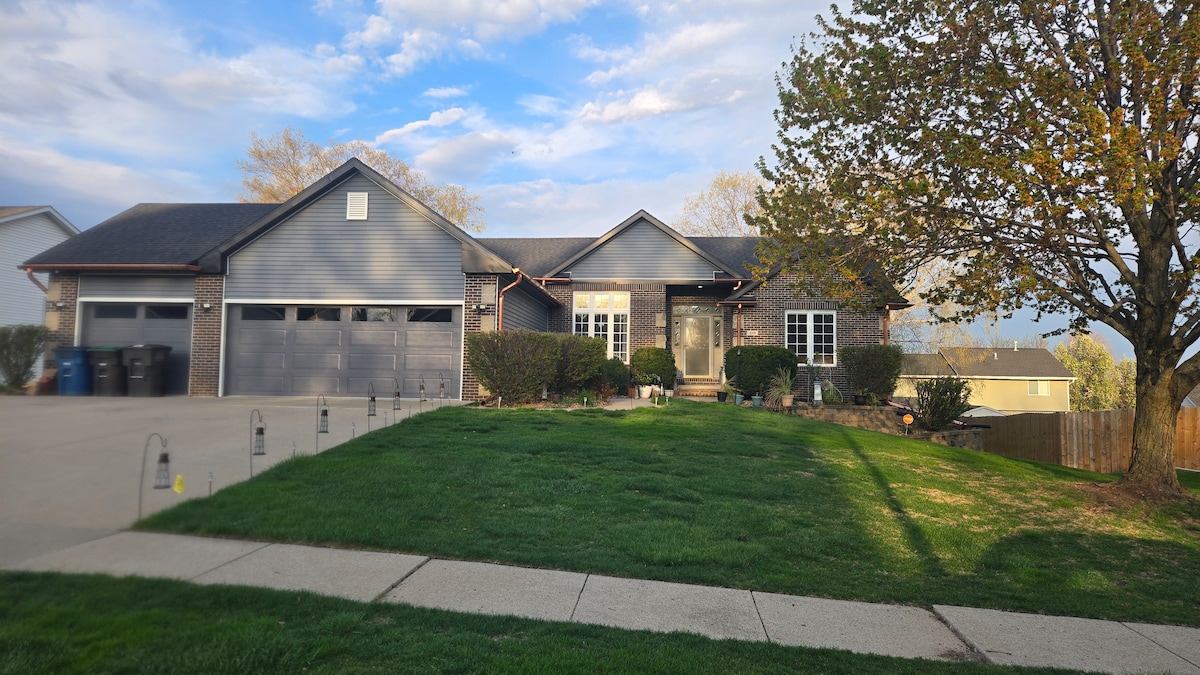 A single-story home features a gray exterior with a centrally located entrance. A manicured lawn is visible in front, along with landscaping elements and a large driveway leading to two garage doors. Trees provide shade, and a clear sky can be seen above.