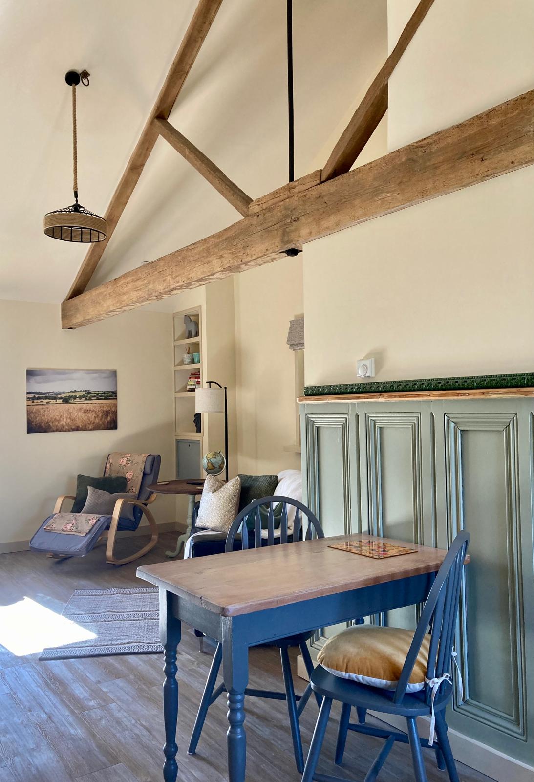The open-plan living area features exposed wooden beams and natural light. A rocking chair and a sofa are positioned near a small dining table with two chairs. A decorative rug adds warmth and comfort, complemented by artwork depicting a tranquil landscape.