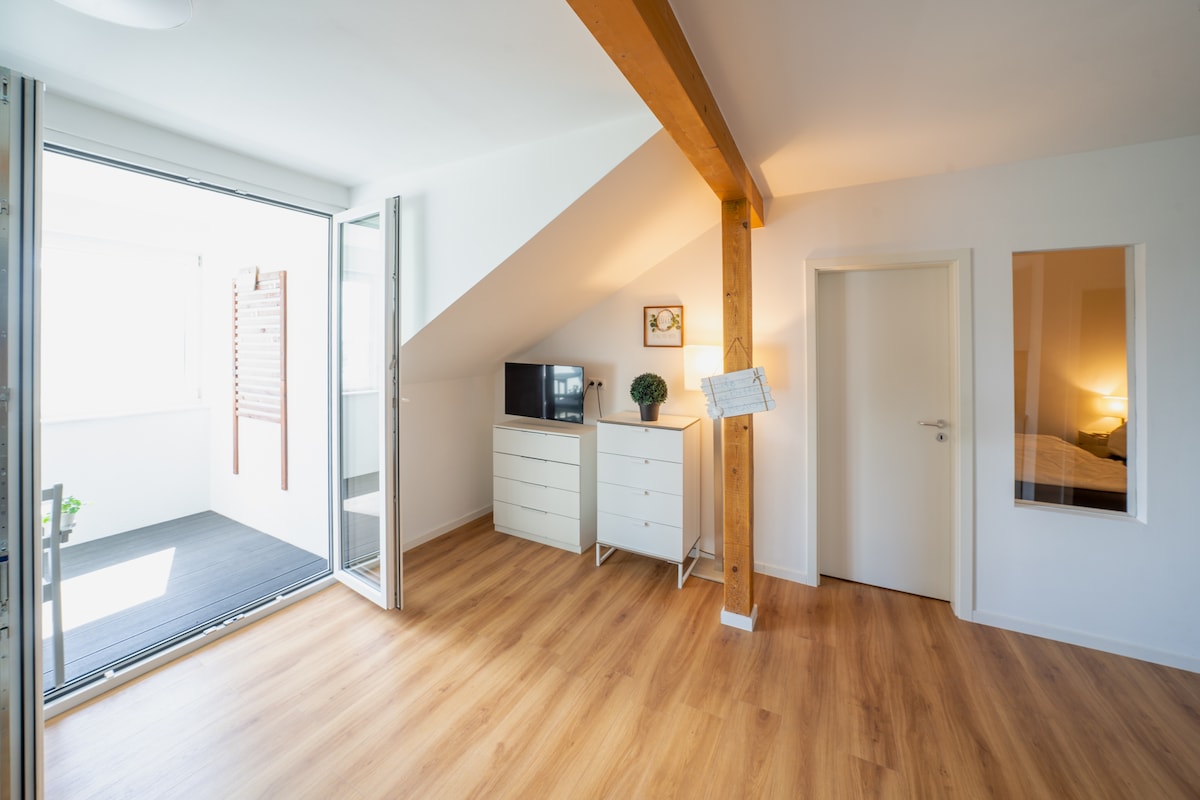 A light-filled interior features a spacious area with wooden flooring and a sloped ceiling. A compact TV stand is positioned alongside a white dresser. A door opens to a small enclosed balcony, while another door leads to a sleeping area visible through a frosted glass panel.