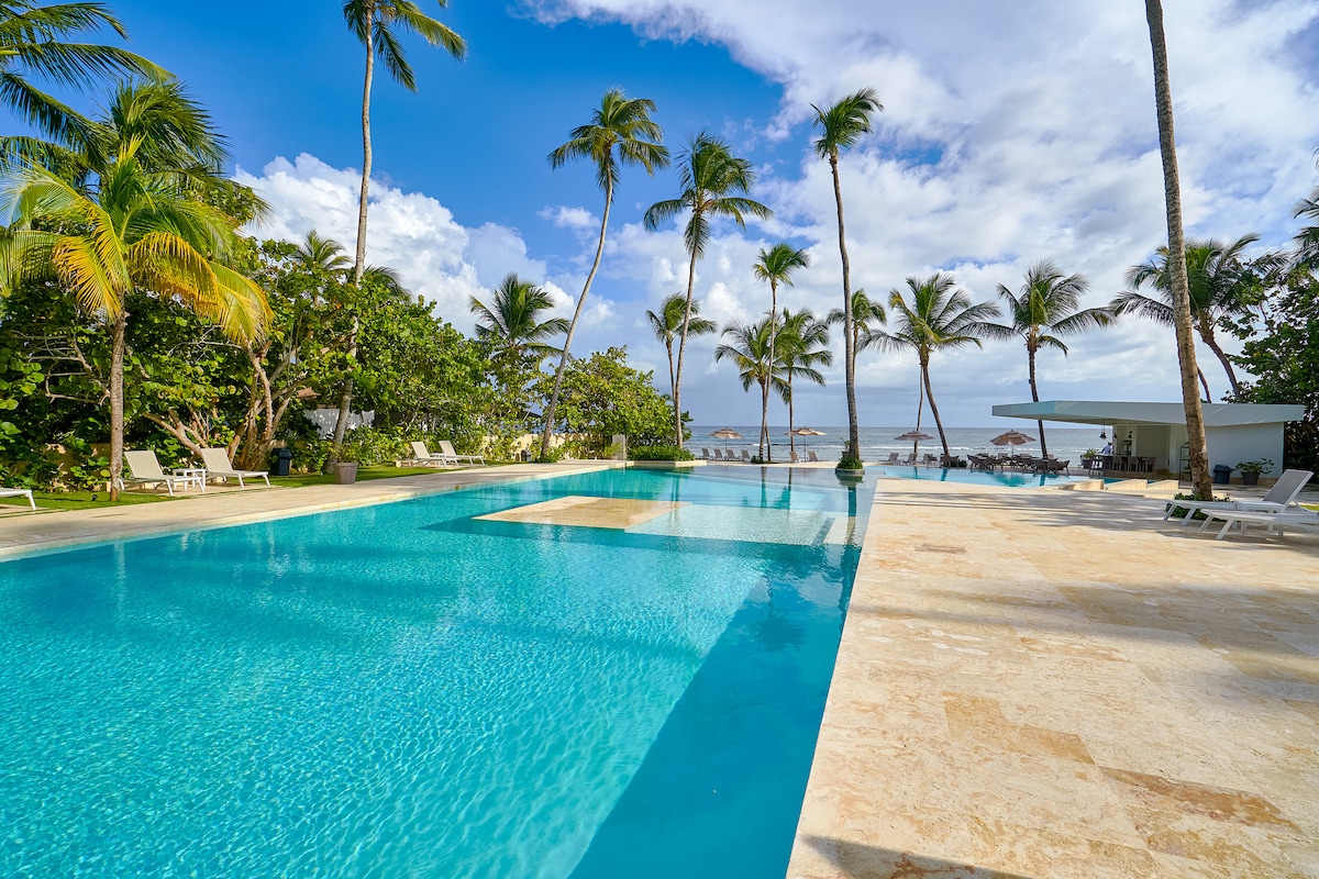 A spacious pool area is surrounded by tall palm trees and tropical plants. The crystal-clear water reflects the blue sky and fluffy white clouds. Lounge chairs are arranged neatly around the pool, offering a relaxing space for guests to enjoy the sunny day.