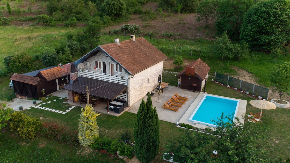 A serene house is surrounded by lush greenery, featuring a well-maintained swimming pool and a shaded patio area. Sun loungers are arranged near the pool, while a wooden structure serves as a barbecue area. A path of stepping stones leads through the garden.