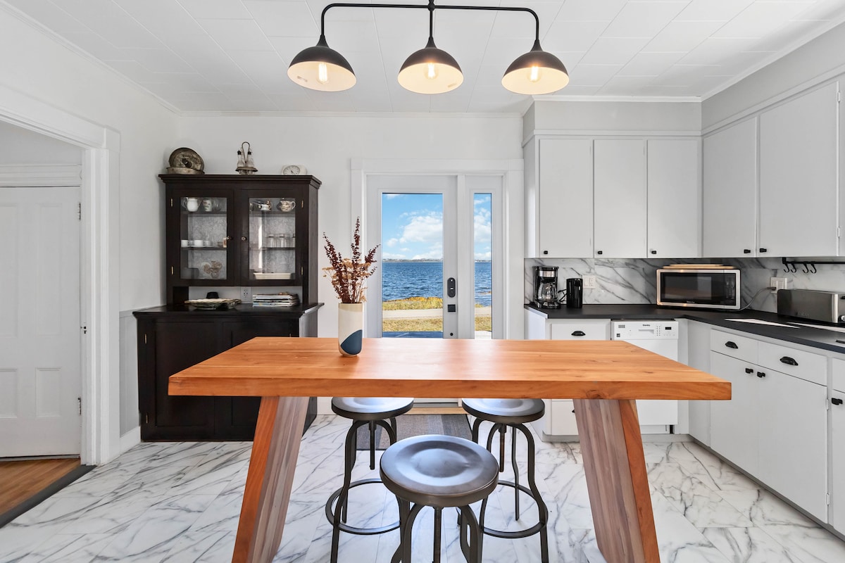 A modern kitchen features a wooden dining table with three stools, surrounded by sleek cabinetry and stainless steel appliances. Natural light floods the space through the glass doors, providing stunning views of the ocean and outdoor scenery.