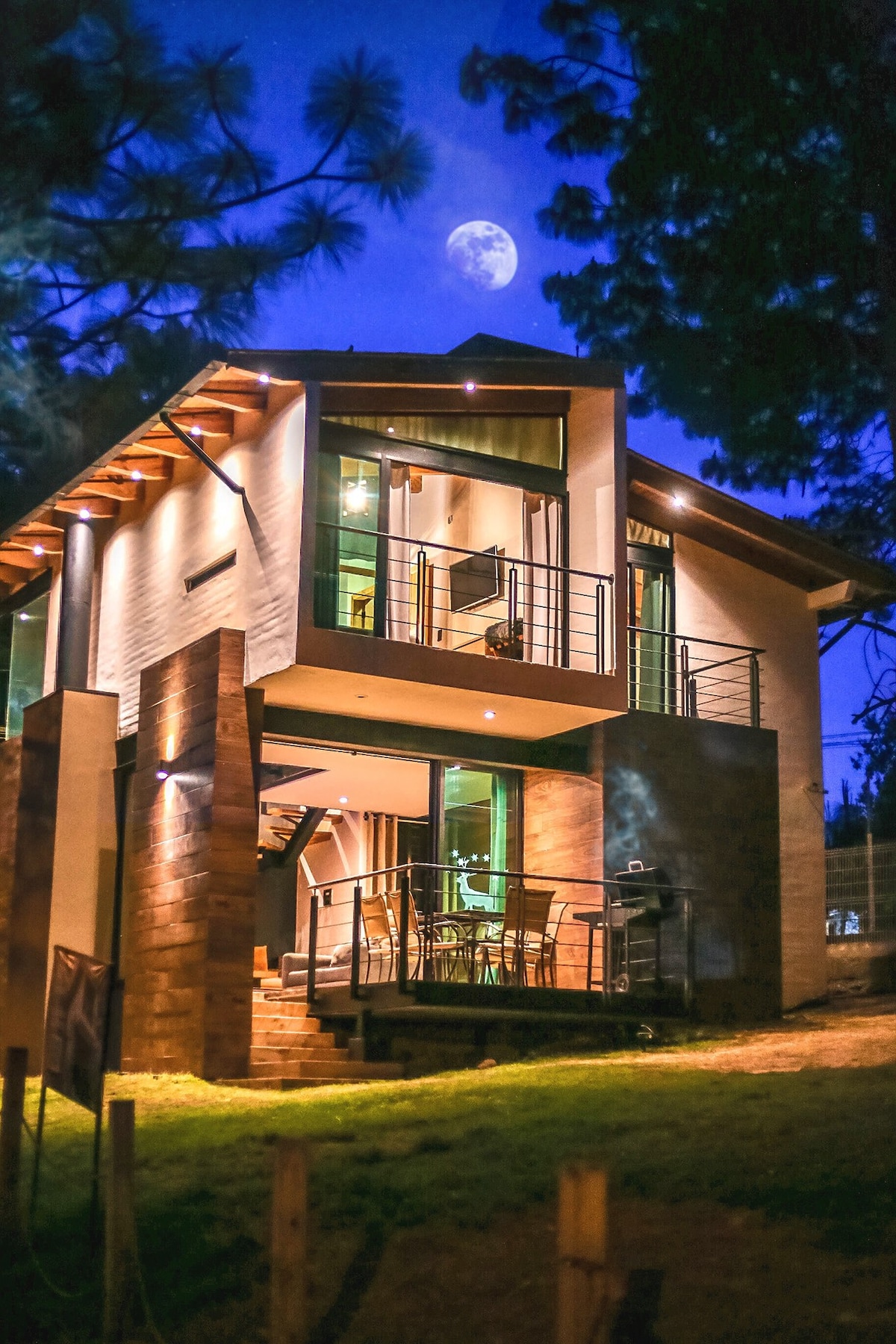 A modern cabin is illuminated at night, showcasing expansive glass windows and a spacious balcony. The structure features a mix of wood and stone elements, surrounded by trees. A full moon is visible in the sky above, adding to the serene atmosphere.