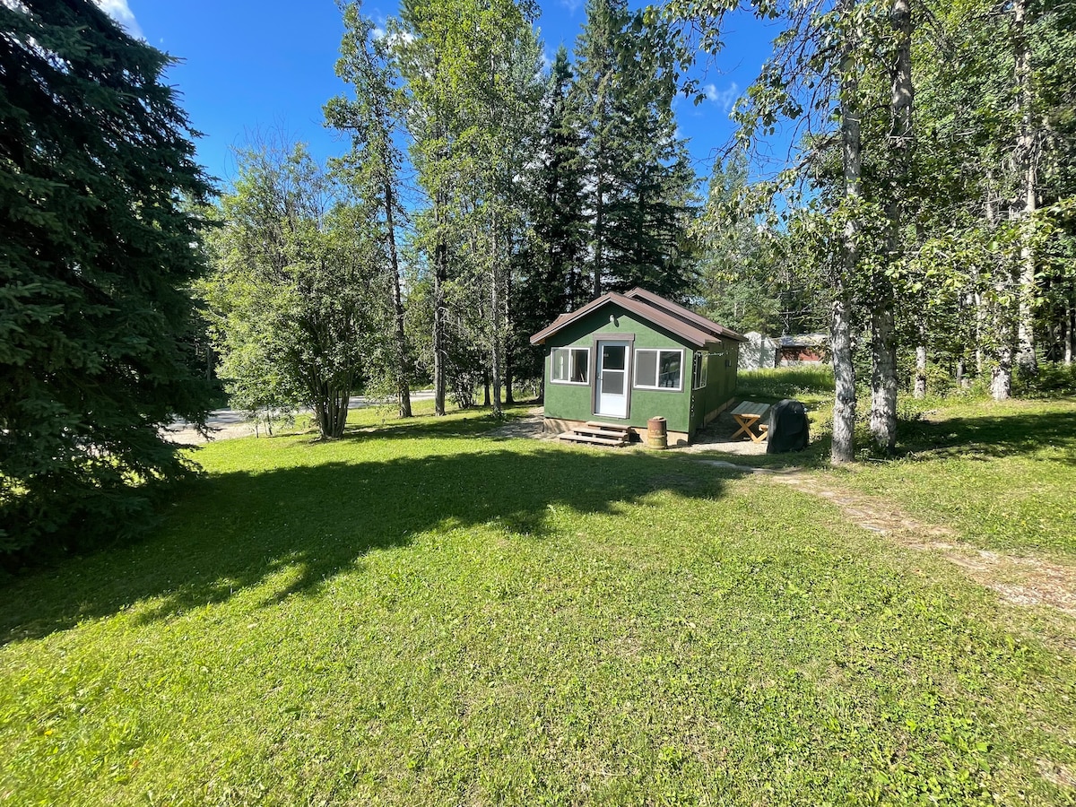 The cabin's exterior is framed by lush greenery and tall trees, providing a sense of tranquility. It features a welcoming porch and large windows, allowing natural light to fill the interior. The surrounding yard is spacious, perfect for outdoor activities and relaxation.