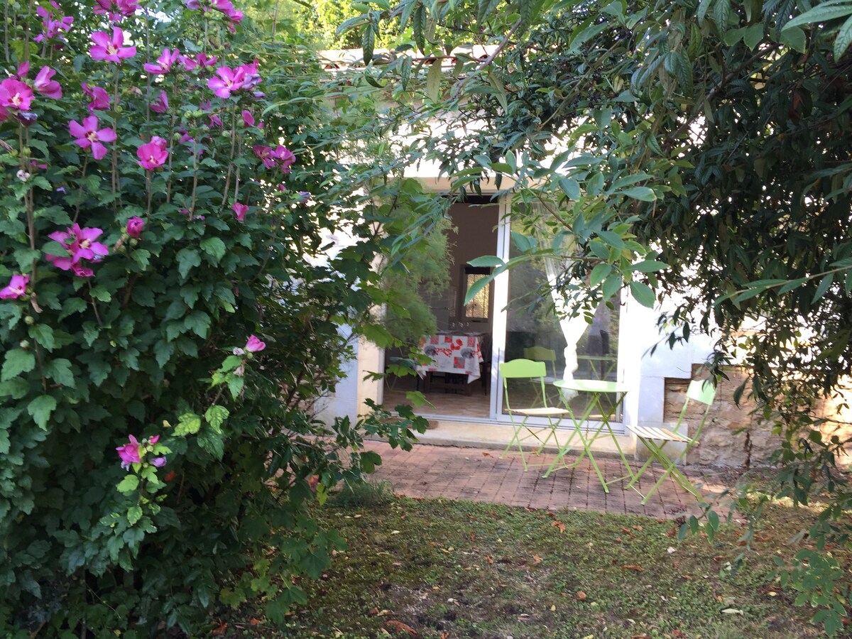 A private terrace is visible through lush greenery and blooming pink flowers. Green outdoor furniture is positioned on the terrace, creating a serene outdoor space. The entrance of the gîte-studio, with large glass doors, invites natural light into the interior.