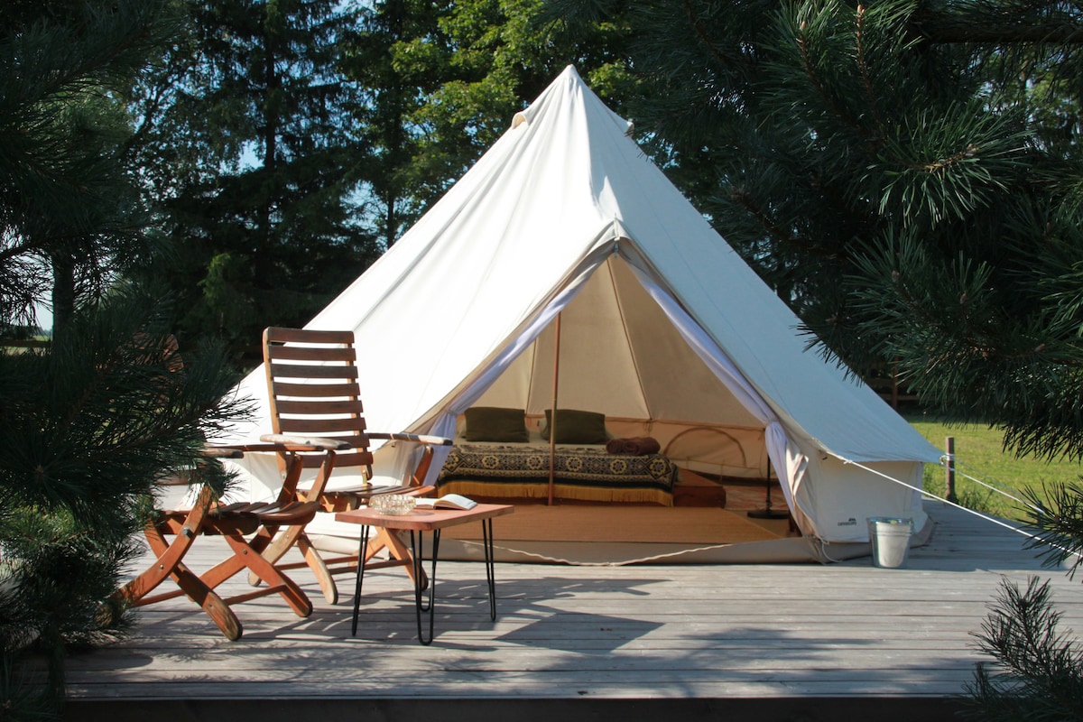 A glamping tent is set on a wooden terrace, surrounded by greenery. Two wooden chairs and a small table offer a relaxing space outside. The tent features an entrance revealing a cozy interior, designed for comfortable outdoor living.