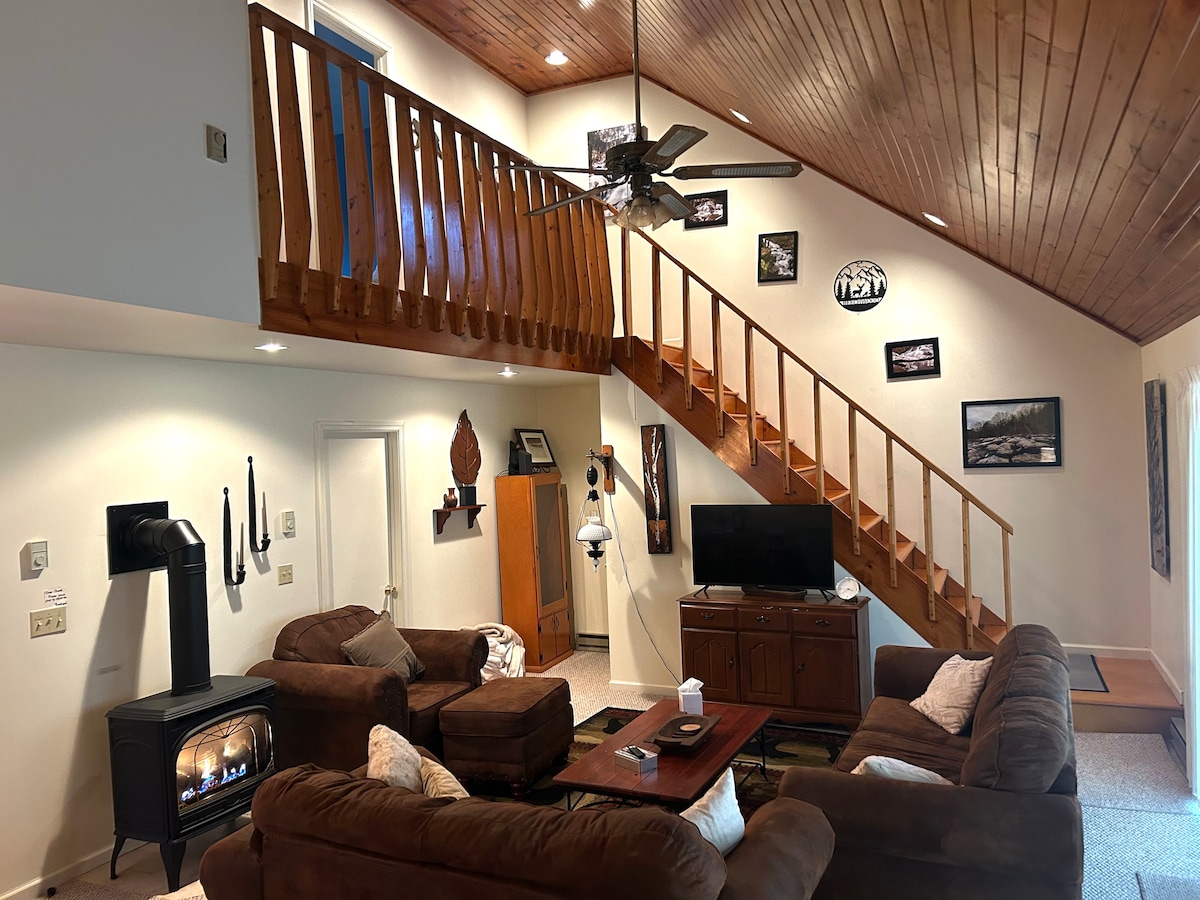 The open living area features a high ceiling with wooden beams and a cozy seating arrangement centered around a fireplace. A staircase leads to the balcony, while various wall art pieces add character to the space. Natural light enhances the inviting atmosphere.