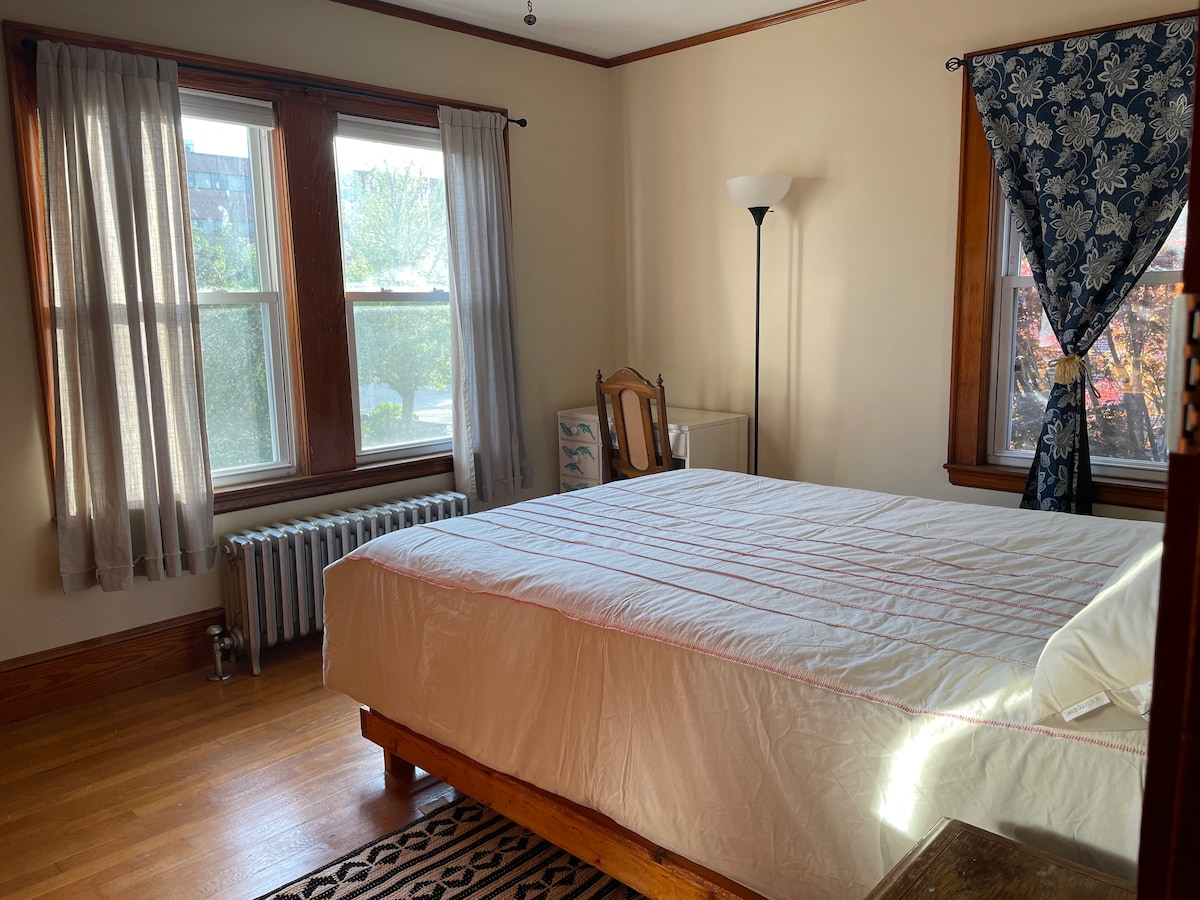 A private bedroom features a spacious bed covered with a light-colored quilt, positioned against a hardwood floor. Two large windows allow natural light to illuminate the room, complemented by light curtains. A lamp stands beside the bed, and a chair is placed nearby.