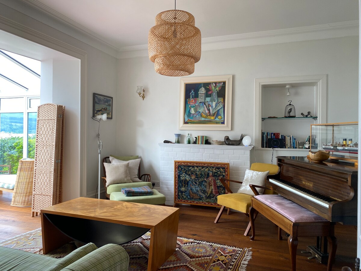 A comfortable sitting area showcases a mix of seating options, including a green sofa, yellow chairs, and a wooden coffee table. A decorative rug lies on the wooden floor, while a grand piano rests against the wall. A fireplace is adorned with colorful artwork, complemented by a stylish light fixture overhead.