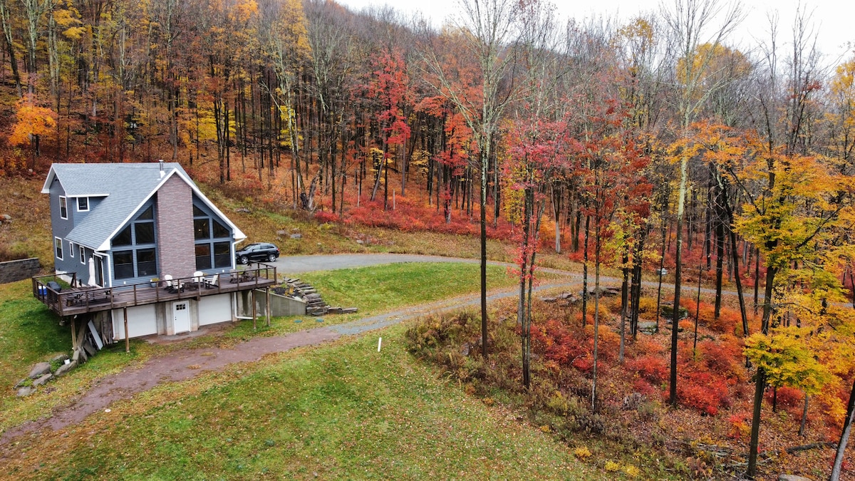 A stylish chalet is situated among vibrant autumn foliage, featuring a wraparound deck that enhances views of the colorful trees. A winding driveway leads to the entrance, and the lush green lawn complements the natural surroundings, creating a serene outdoor setting.