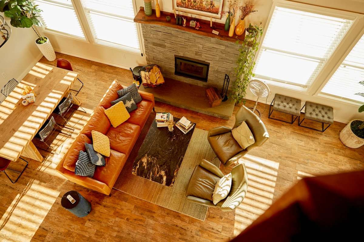 The spacious living area features a warm leather sofa adorned with colorful cushions, positioned adjacent to a natural stone fireplace. A wooden dining table sits nearby, complemented by two armchairs on a decorative area rug. Sunlight streams through large windows, casting patterned shadows across the floor.