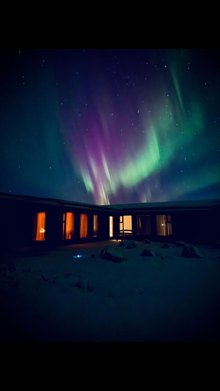 A modern house is illuminated under a vibrant display of the northern lights, with hues of green and purple visible in the night sky. Snow blankets the ground, contrasting with warm light emanating from the windows, creating a serene atmosphere.