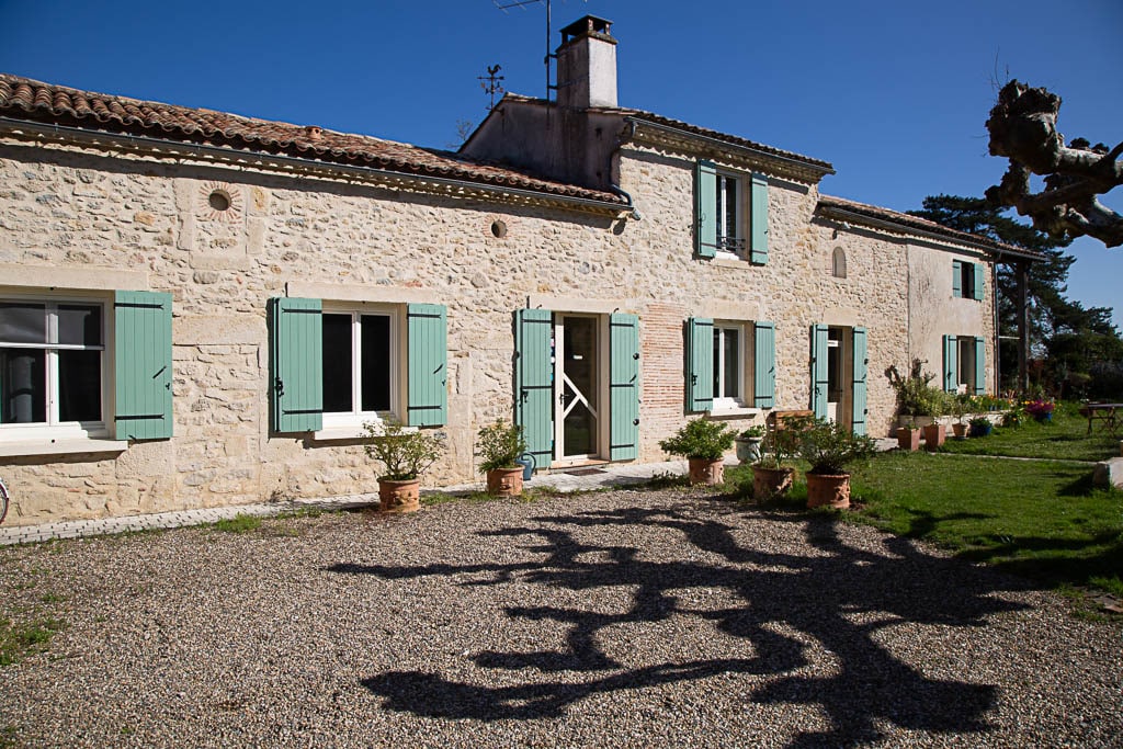 The charming stone exterior of the house is complemented by light green shutters and a gravel pathway. Flowering pots line the entrance, enhancing the home's natural setting. A clear blue sky provides a bright backdrop, highlighting the peaceful ambiance of the property.