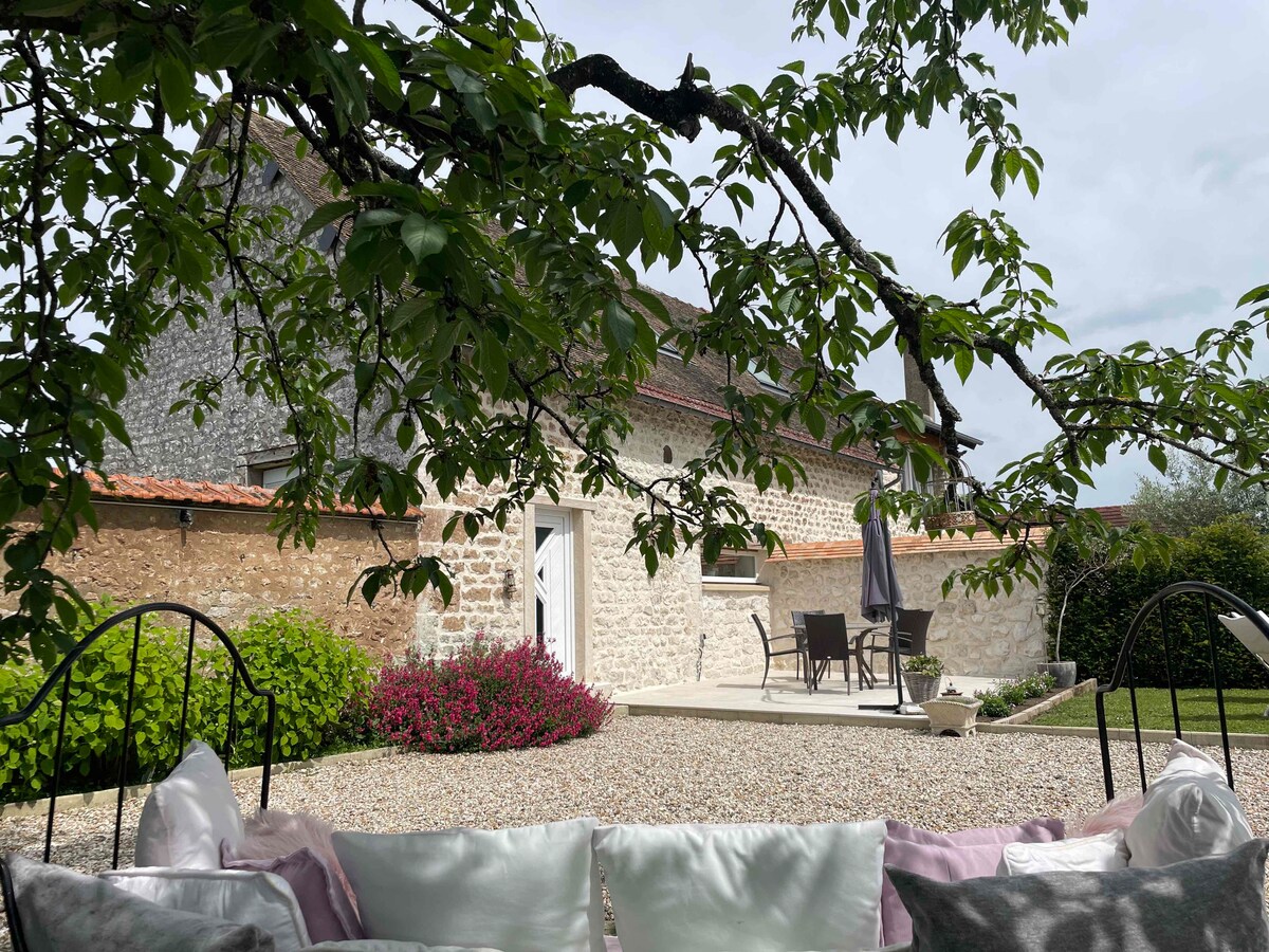 A view of the renovated gîte showcases a stone facade, surrounded by lush greenery and flowering shrubs. An outdoor patio with seating is visible, shaded by a tree. The inviting garden space features a mix of gravel and grass, enhancing the tranquil atmosphere.