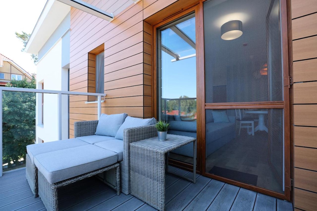A serene balcony space is featured, furnished with a gray wicker lounge chair and a small table. A potted plant is placed on the table. Glass sliding doors provide access to the interior, while a partial view of the surrounding area is visible.