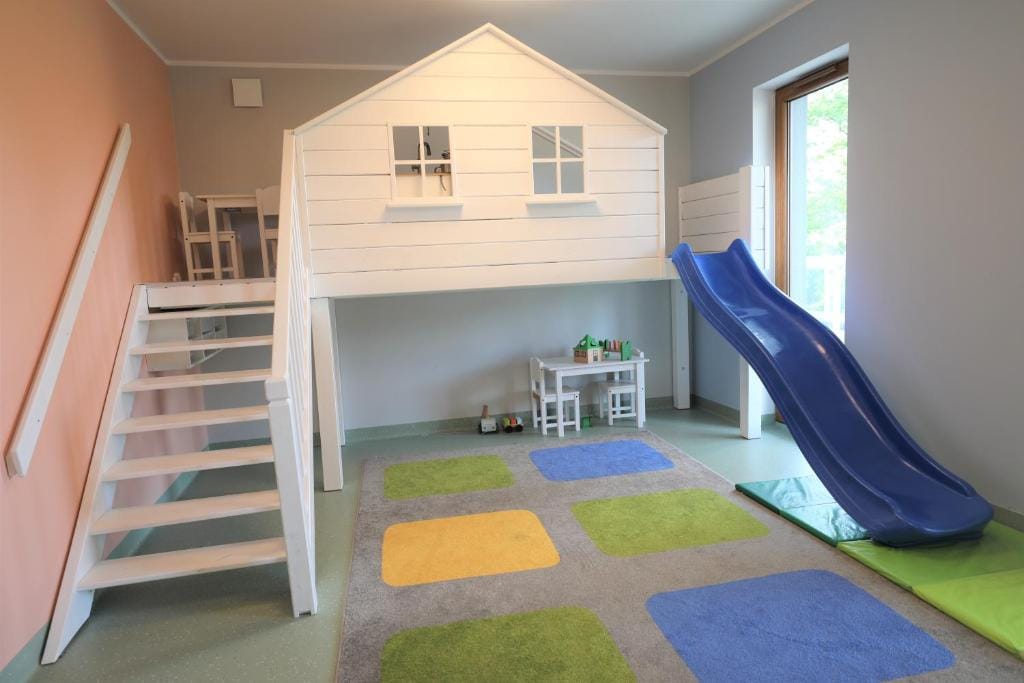 A children's playroom is featured, with a white, house-shaped play structure that includes a slide and two windows. A small table with chairs is positioned nearby. Colorful rugs in blue, yellow, and green enhance the playful environment, complemented by soft lighting from the window.