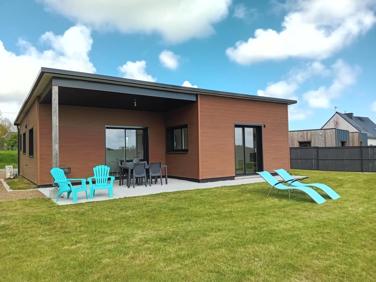 A modern wooden house features large windows and a spacious patio area. Colorful lounge chairs and a dining set are arranged on the patio, surrounded by an open grassy space. The building showcases a simple, clean design with a muted color palette.