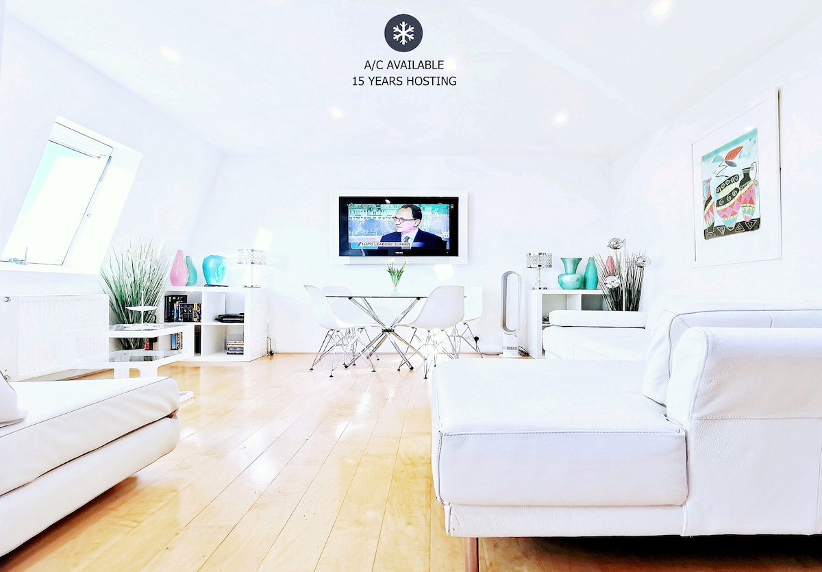 A bright living area is highlighted by polished hardwood flooring and contemporary white furnishings. A sleek round dining table is positioned centrally, with modern decor and green plants adding a touch of freshness. A flat-screen television is mounted on the wall, creating a cozy atmosphere.
