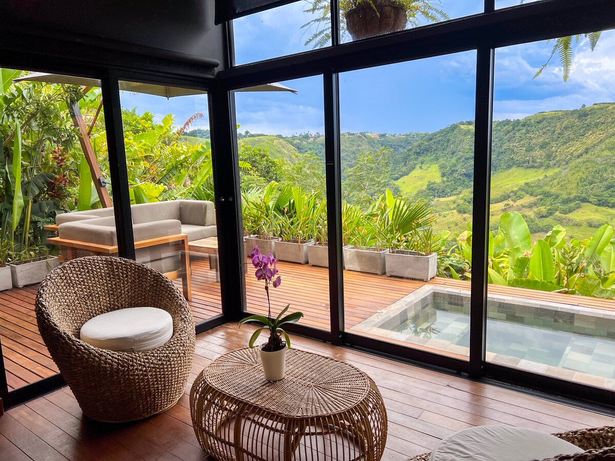 A cozy seating area features natural woven furniture and a small potted plant with purple flowers. Expansive glass doors offer a view of lush green hills and a wooden deck with a hot tub, surrounded by tropical plants and trees.