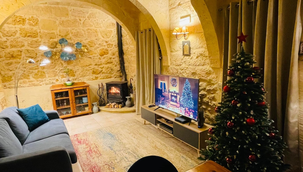 A cozy living area features stone walls and an inviting wood-burning fireplace. A green sofa is positioned in front of a large television. A decorative Christmas tree stands in the corner, and soft lighting creates a warm ambiance. Curtains frame the windows, enhancing the intimate setting.