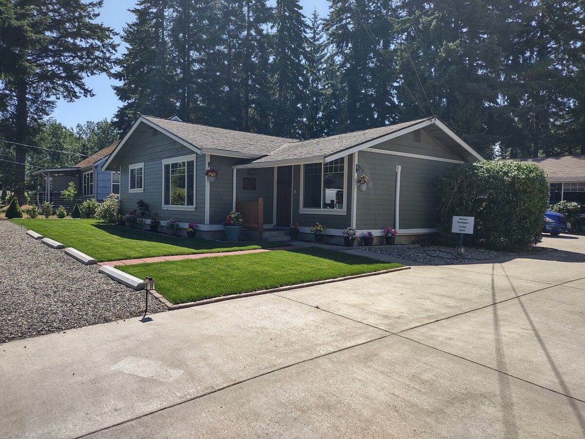 A charming single-story home is situated within a quiet residential area. The exterior features a well-maintained lawn and garden, with flower pots lining the entrance. Large windows allow natural light to brighten the interior, while tall trees provide a serene backdrop.