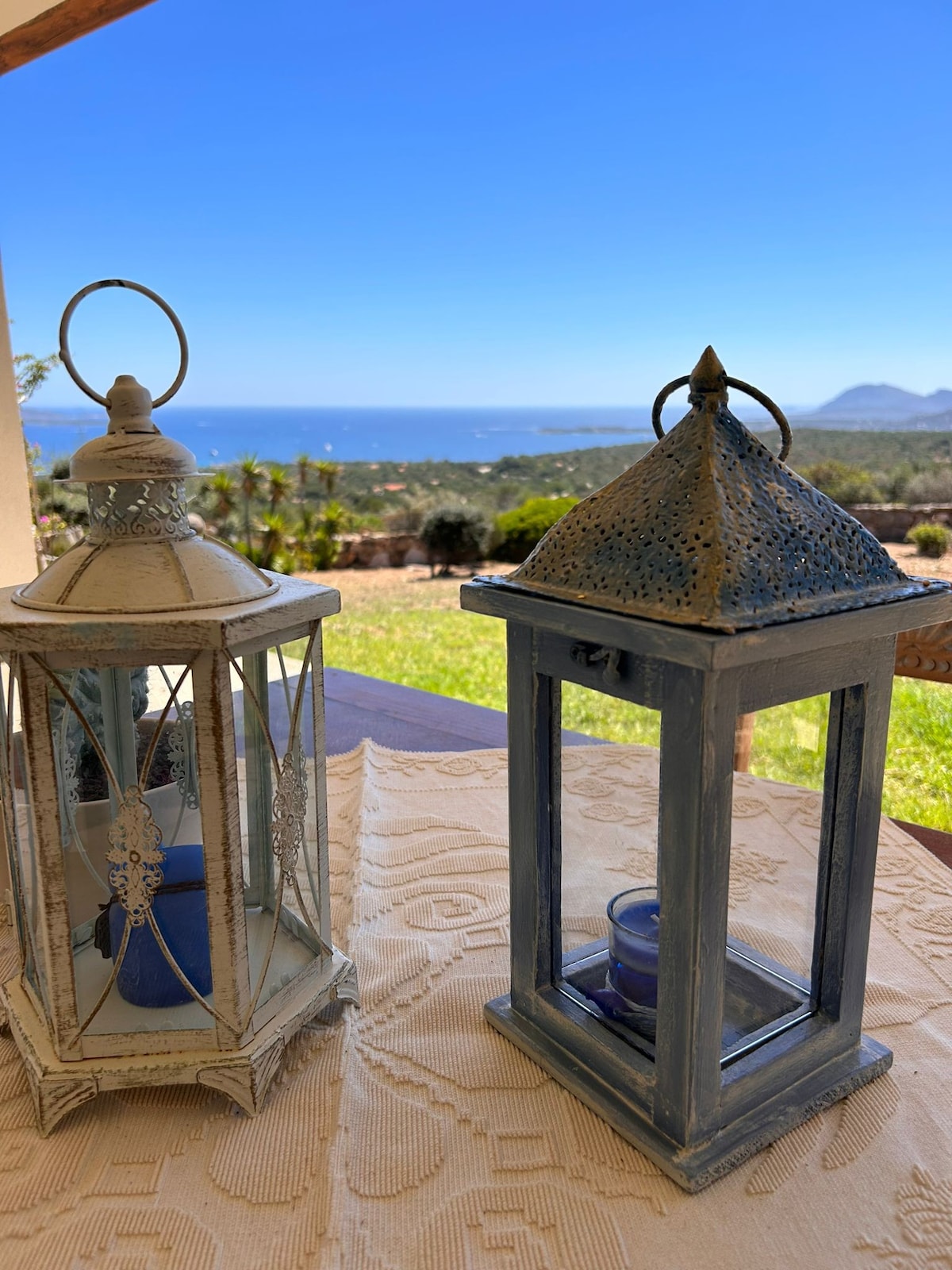 Two decorative lanterns are displayed on a textured tablecloth, set against a backdrop of a panoramic seascape. The vibrant blue sky meets the water in the distance, while lush greenery is visible in the foreground, creating a serene outdoor atmosphere.