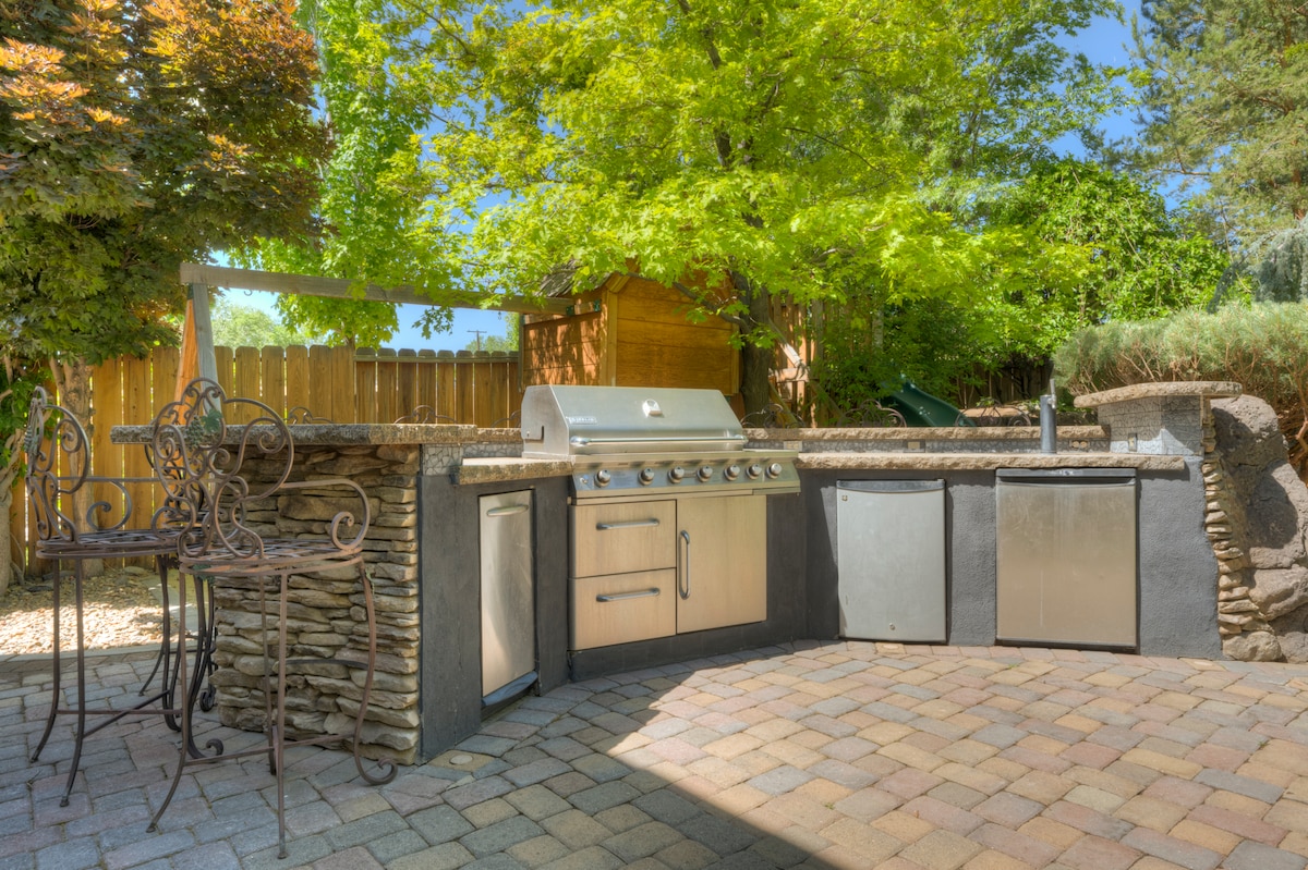 An outdoor grilling area is presented, featuring a stainless steel grill and storage spaces integrated into a stone structure. Two metal stools are positioned at the bar, surrounded by lush greenery and a wooden fence that enhances privacy.