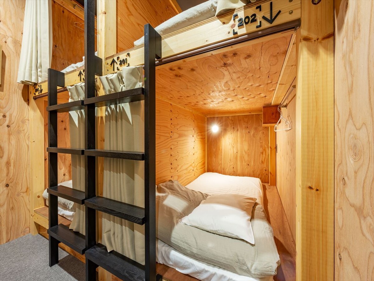 A compact dormitory room features wooden walls and ceiling, creating a natural ambiance. A bunk bed is visible, with a spacious bed below and a sleeping area above, softly lit by a warm light. Curtains provide privacy for guests.