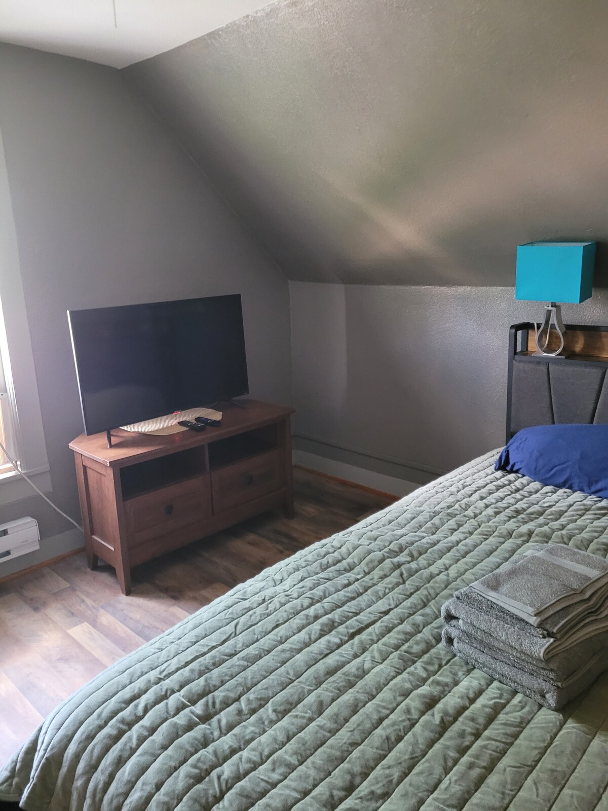 A private room features a spacious bed with a neatly arranged set of towels. A large TV is positioned on a wooden dresser, and a window allows natural light to enter the room, complementing the muted wall colors.