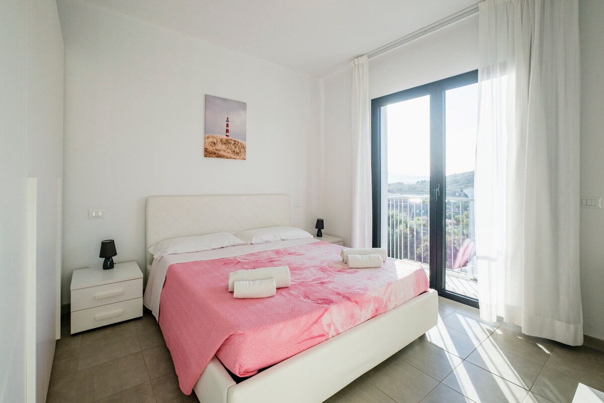 A spacious bedroom is highlighted by large glass doors opening to a balcony, allowing natural light to fill the room. A large bed with crisp linens is centered on the floor, accompanied by nightstands on either side. Soft, light-colored curtains frame the windows.