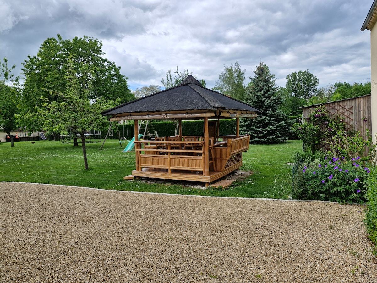 An open wooden gazebo is situated on a grassy area, surrounded by trees and colorful flowers. The gazebo features a large table with built-in benches, providing ample space for dining. Nearby, a swing set is visible, enhancing the outdoor leisure space.