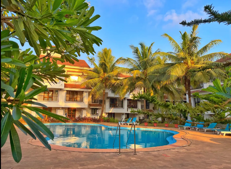 A swimming pool is framed by lush greenery and palm trees, providing a refreshing setting. Sun loungers are arranged around the pool, emphasizing a relaxed ambiance. The building's balconies are visible in the background, enhancing the serene atmosphere of this getaway.