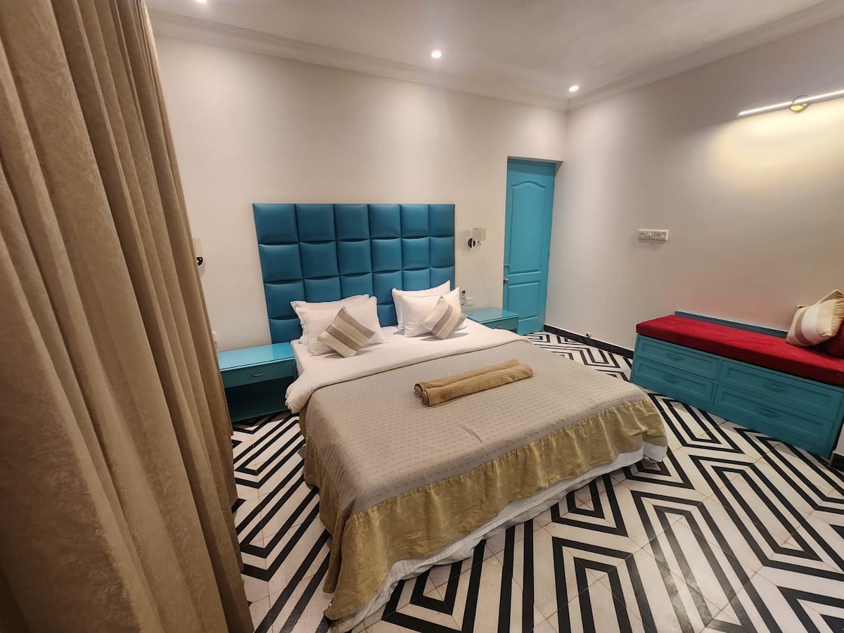 The cozy bedroom features a spacious bed with decorative pillows, flanked by a striking blue headboard. Light-colored bedding contrasts with the geometric patterned floor. A colorful seating area and a door leading to additional spaces are visible in the softly lit room.