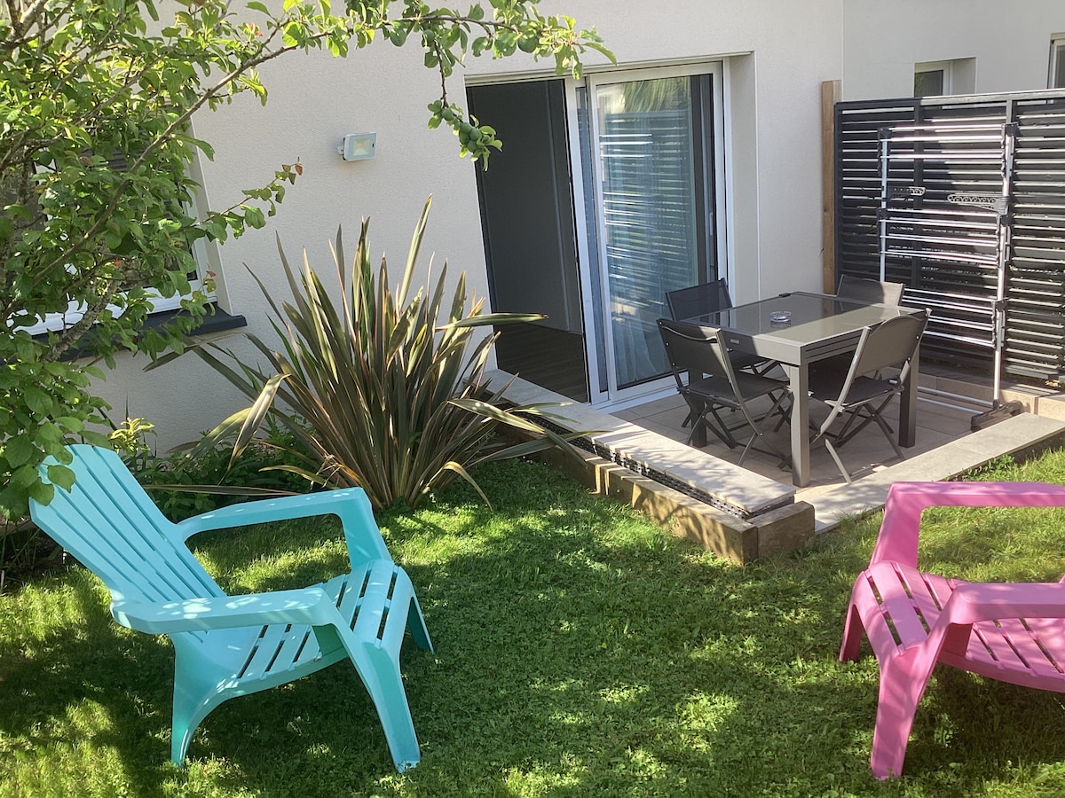 A cozy outdoor space features a small patio area with a table and chairs, surrounded by greenery. Colorful adirondack chairs provide additional seating, while a tall plant adds natural depth to the setting. A sliding glass door offers access to the interior.