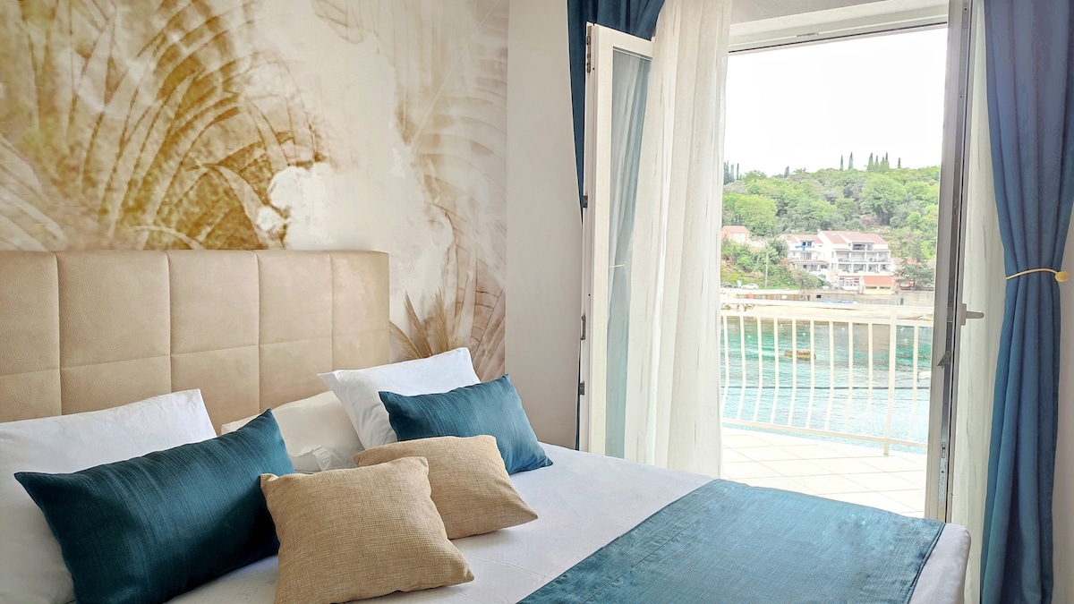 A comfortable bedroom features a king-size bed with crisp white linens and accented by teal decorative pillows. Natural light enters through an open door leading to a private balcony, offering tranquil views of Molunat Bay and greenery. A soft, neutral color palette enhances the serene ambiance.