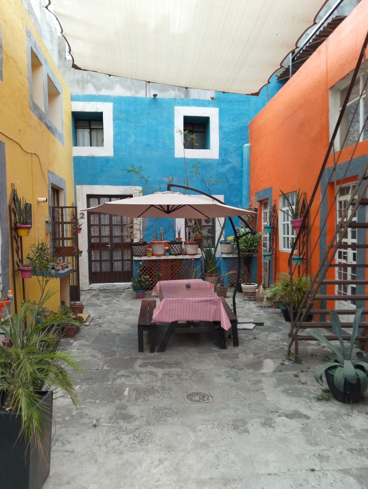A vibrant courtyard features colorful walls in shades of blue and orange, with a shaded area supported by an umbrella over a wooden table. Potted plants and decorative elements add a lively touch, while the stone floor provides a rustic ambiance for relaxation.