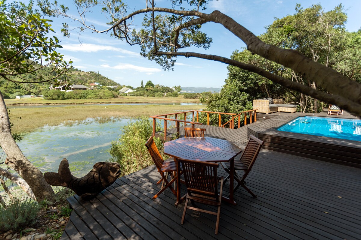 A wooden deck offers outdoor seating around a round table, surrounded by lush greenery. A private swimming pool is visible nearby, with the tranquil Knysna Salt River flowing in the background, framed by trees and gentle hills.