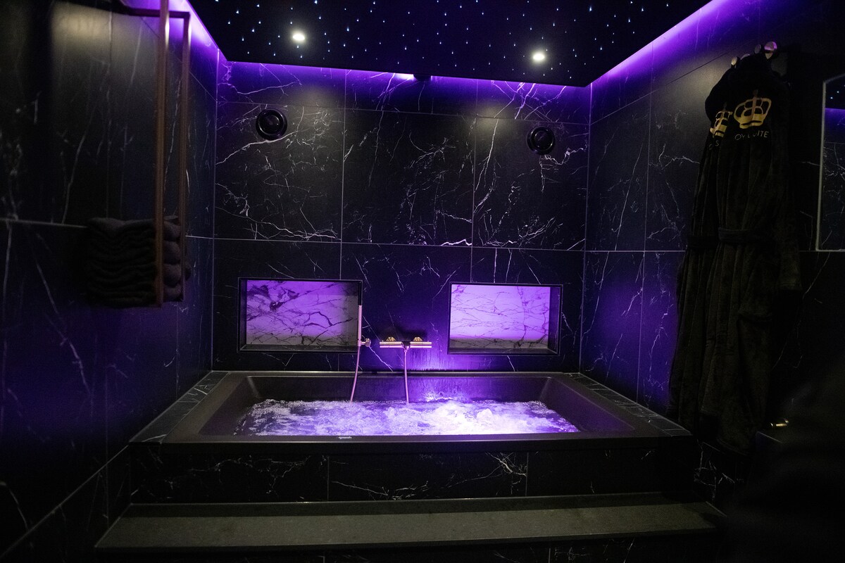 A luxurious bathroom features a bubble bath surrounded by sleek black marble walls. Soft purple lighting enhances the space, while a starlit ceiling creates a serene atmosphere. Two windows showcase marble detailing, and plush robes hang nearby, adding comfort to the setting.