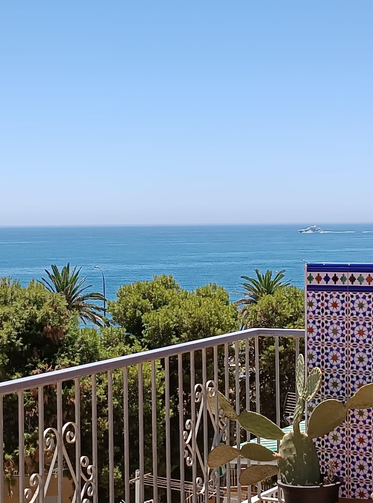 A panoramic view of the sea is presented from the terrace, framed by lush greenery and ornamental tiles. The clear blue waters extend to the horizon, with a boat visible in the distance. The scene reflects the tranquil coastal ambiance.