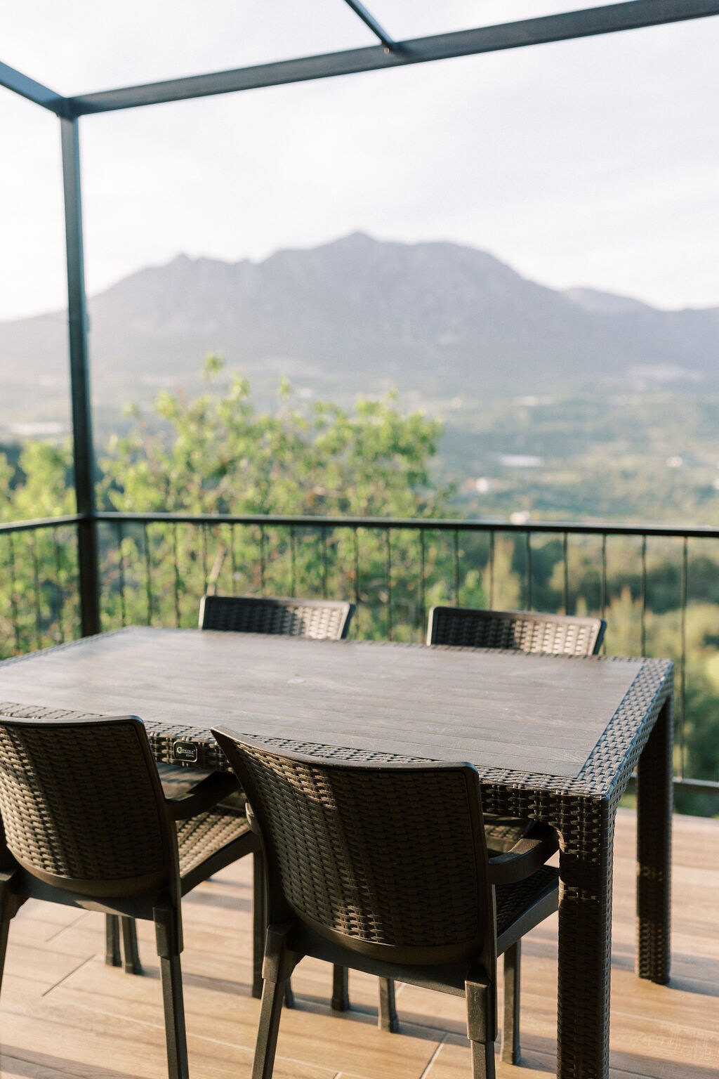 An outdoor dining area features a large rectangular table surrounded by four wicker chairs. The setting offers views of lush green surroundings and distant mountains, framed by a protective canopy that allows for a comfortable dining experience.