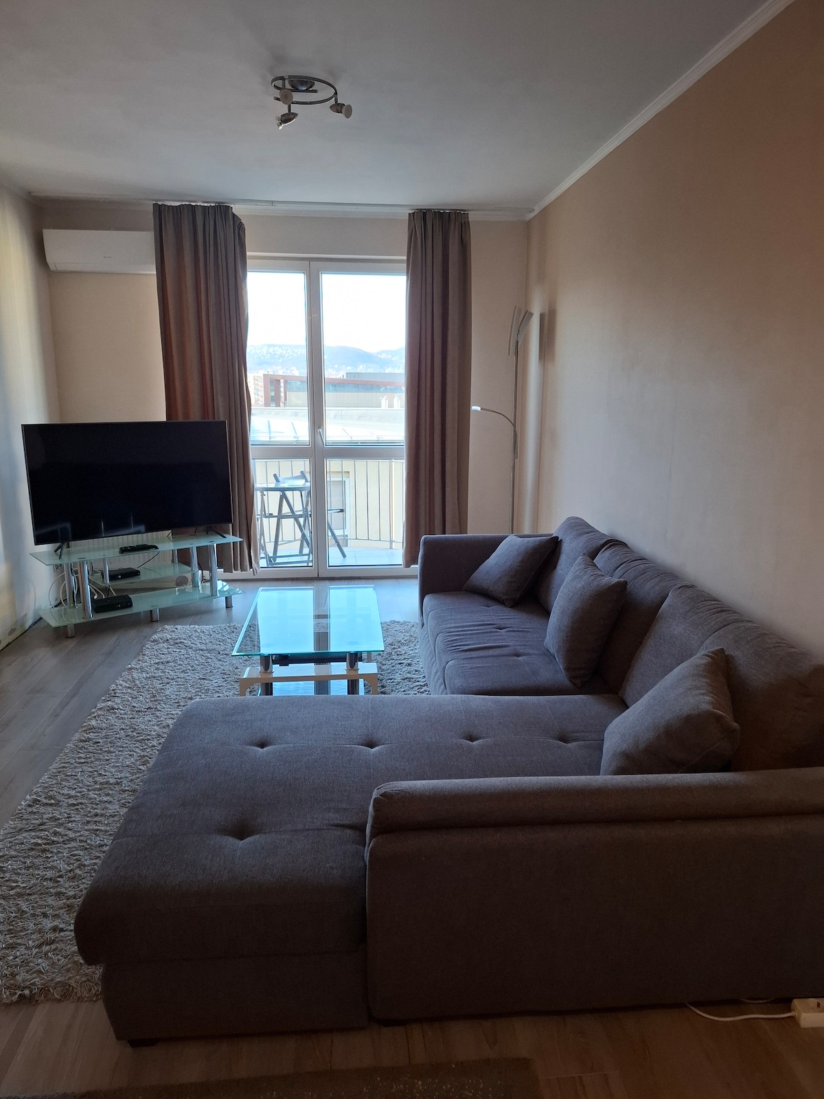 A modern living area features a spacious sectional sofa and a glass coffee table on a light rug. A flat-screen TV is positioned on a stand, and large windows with light curtains provide natural light and a view of the outside.